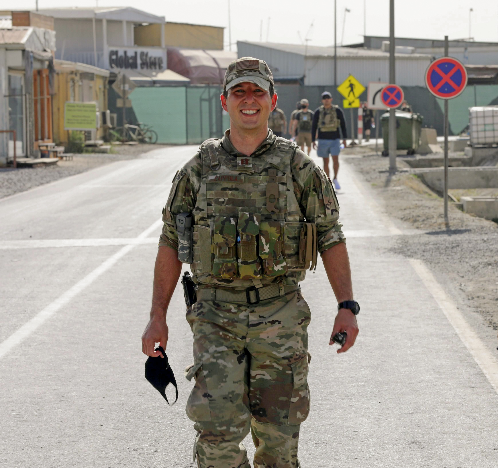 Then-Captain Joseph Cappola poses in uniform in Afghanistan with individuals walking behind him as part of the 9/11 Memorial Ruck March he organized.